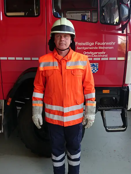 Feuerwehrmann in orangefarbener Einsatzkleidung mit Helm vor einem Löschfahrzeug